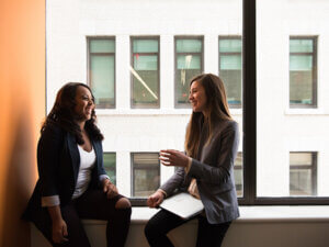 photo of two professional women sitting on a windowsill in a library or office building and talking