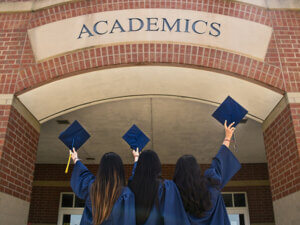 photo of three people in graduation gowns holding up their caps in front of a brick building with the sign "Academics" across the top